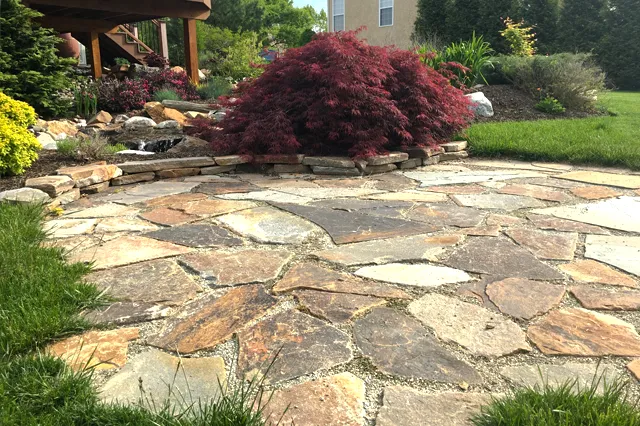 Irregular dry-laid flagstone patio with stone retaining wall edging, red Japanese maple, and mixed plantings with water feature in background