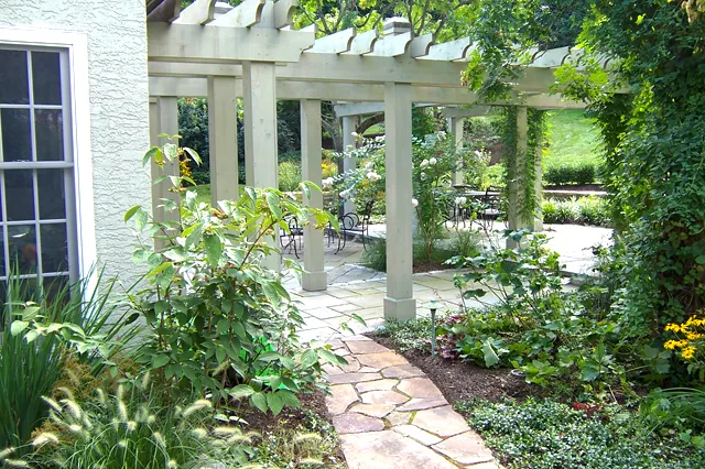 White painted pergola with climbing vines and flagstone path leading to a patio dining area, surrounded by lush mixed garden borders