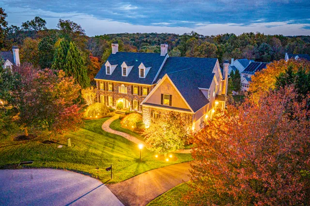 Aerial dusk view of a stone and shingle manor home with professional landscape uplighting on trees and path lighting along the driveway, surrounded by fall foliage
