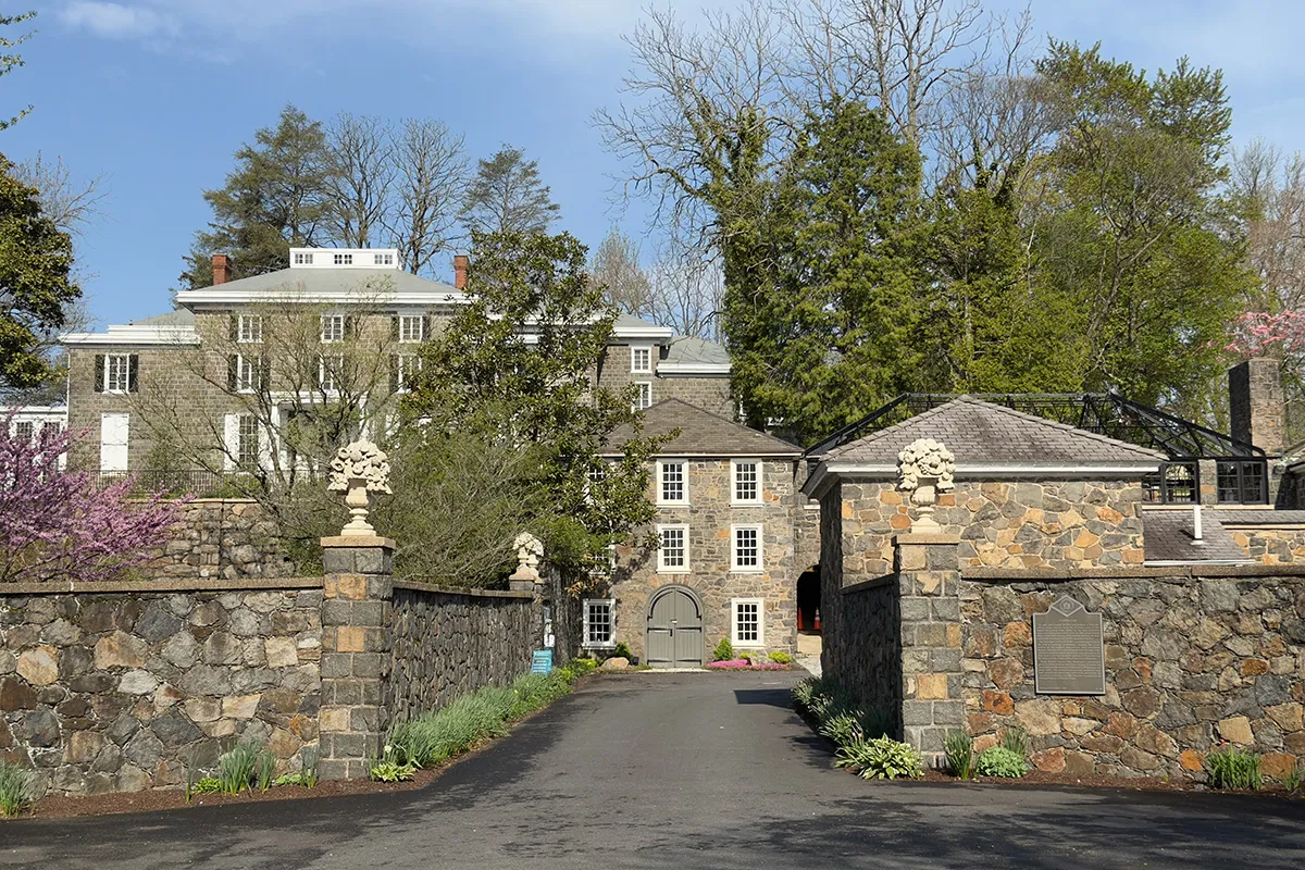 Stone gate entrance to Gibraltar Estate in Wilmington, Delaware, with historic fieldstone boundary walls, ornamental finial pillars, and a driveway leading to the stone manor house amid spring flowering trees