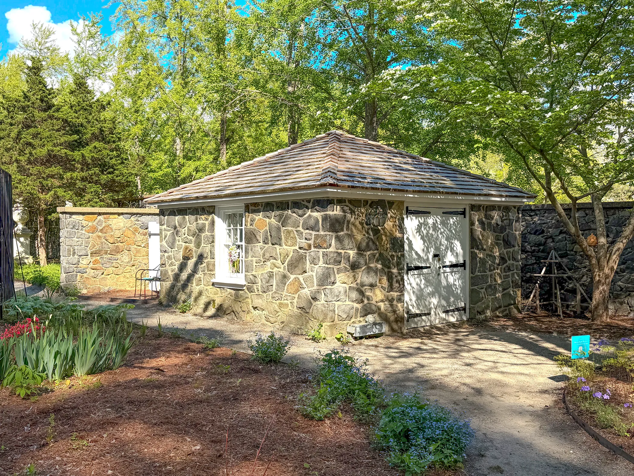 Historic Brandywine granite gardener's cottage with hip roof, white trim doors and window, spring garden beds of tulips and iris in foreground, stone perimeter wall behind, Gibraltar Estate, Wilmington, DE