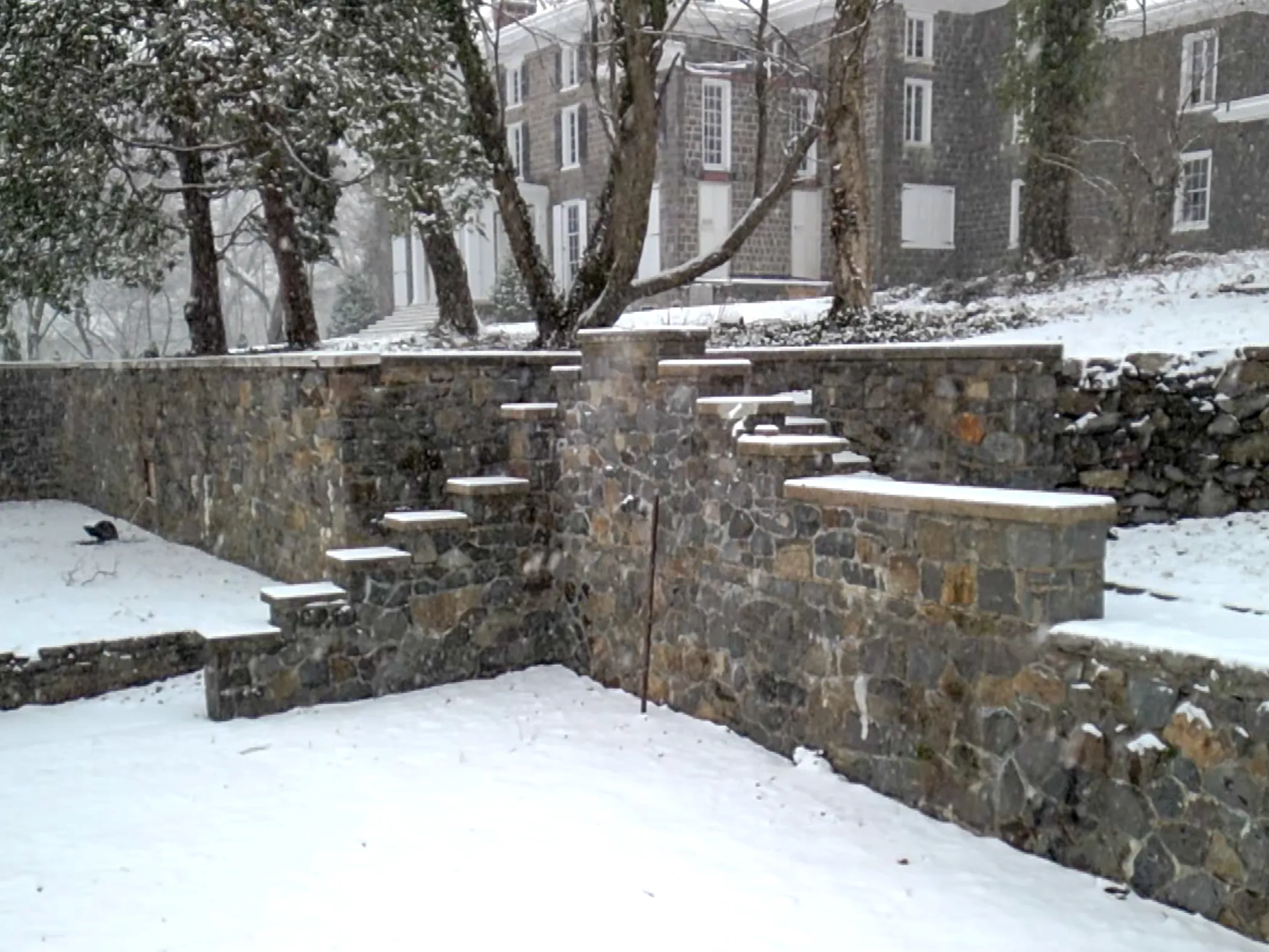 Terraced Brandywine granite retaining walls with stepped stone coping caps dusted in snow, Gibraltar mansion visible through bare trees in background, Wilmington, DE