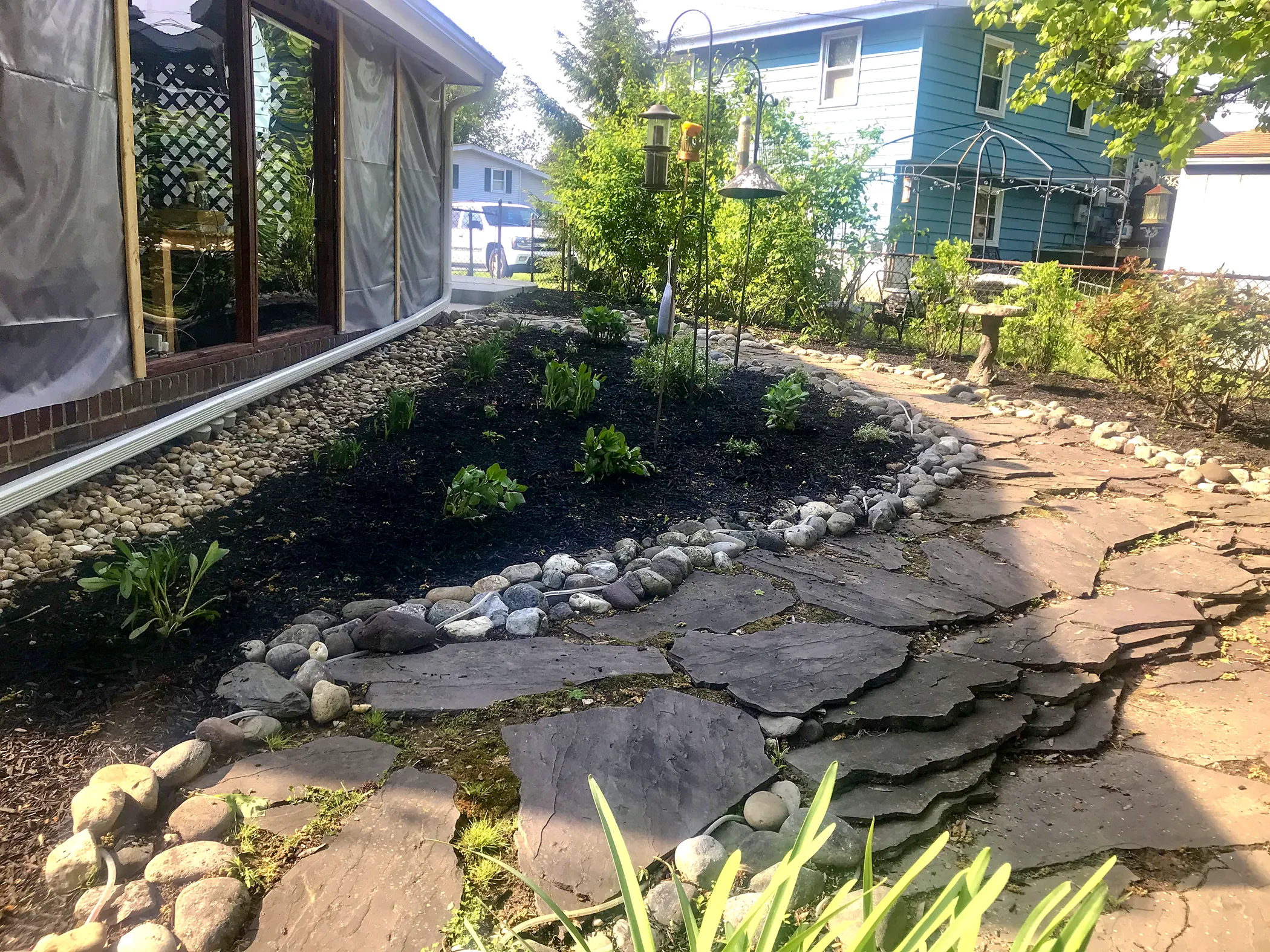 Newly installed backyard wildlife habitat garden with natural flagstone path, river rock edging, perennial planting beds, and bird feeder stations adjacent to a screened porch