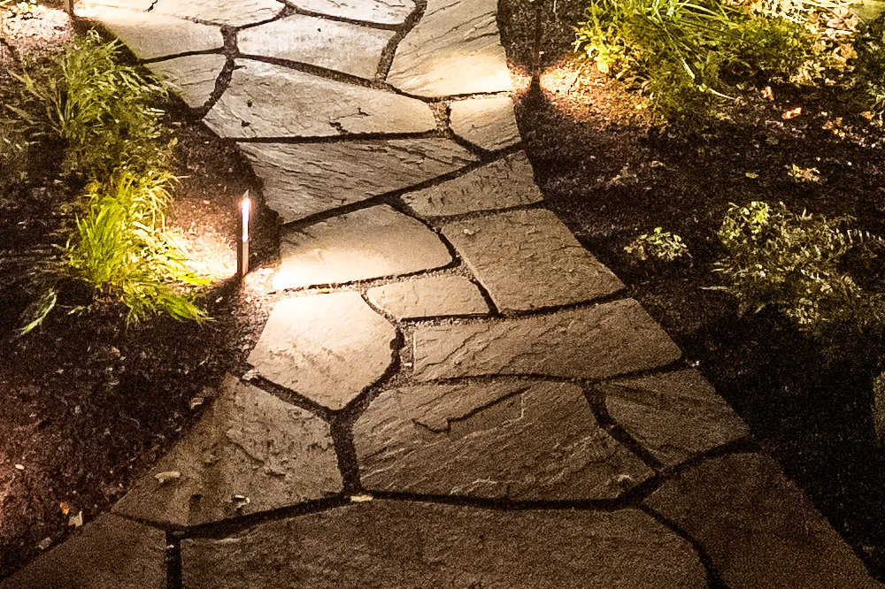 Low-voltage LED bollard path light set beside an irregular flagstone walk with lit ornamental grasses — landscape lighting installation by Eastern Horticultural Services