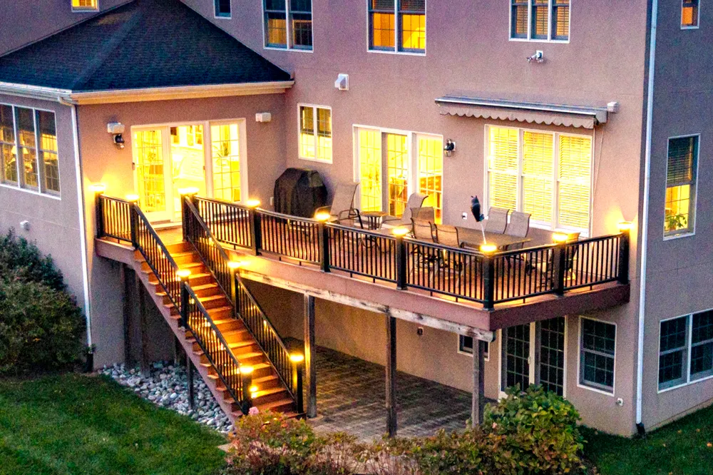 Rear deck at dusk with step riser lighting, rail post cap lights, and interior light glowing through French doors — landscape lighting by Eastern Horticultural Services, Kennett Square, PA