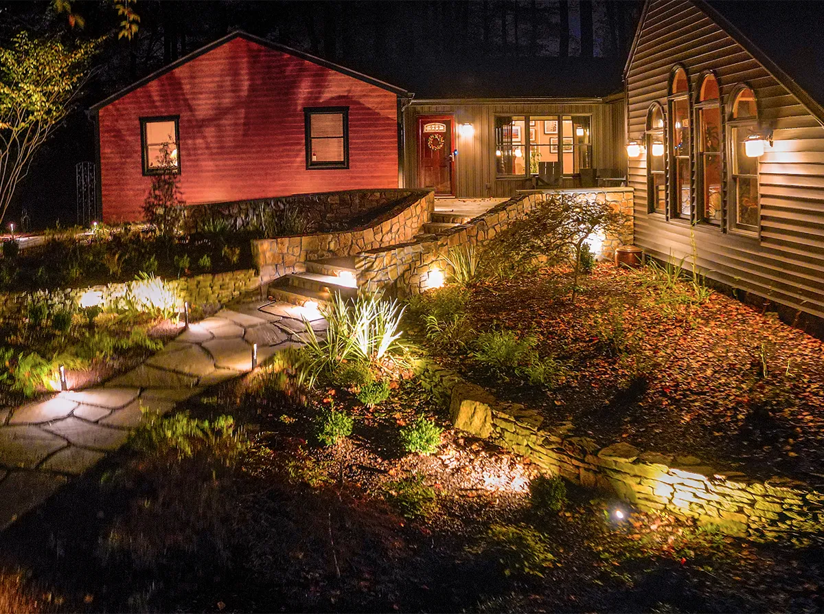 Landscape lighting installation at dusk — flagstone walkway with bollard path lights and uplights illuminating stone retaining walls, steps, and planted beds leading to a residential entry